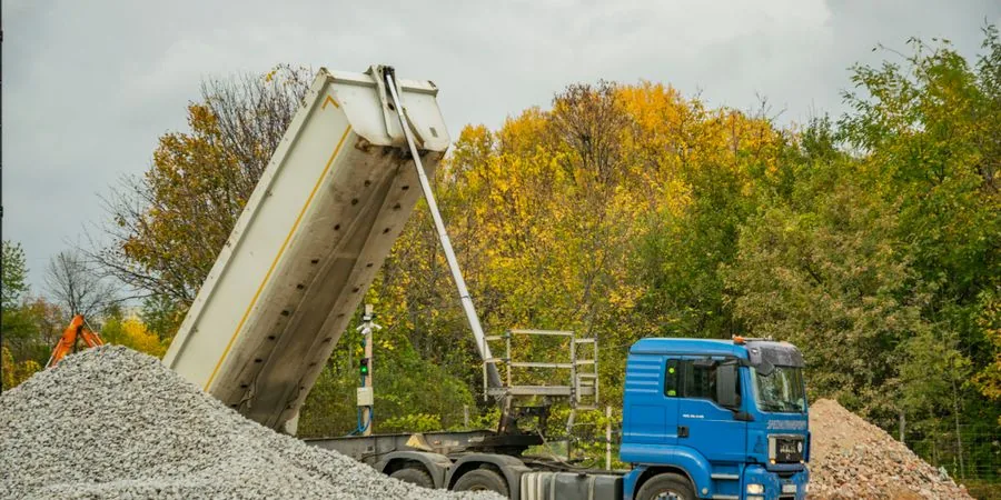 Blue dump truck unloading gravel at an outdoor construction site — the tip-and-discharge action that a self-loading mini dumper replicates at compact scale without a separate loader