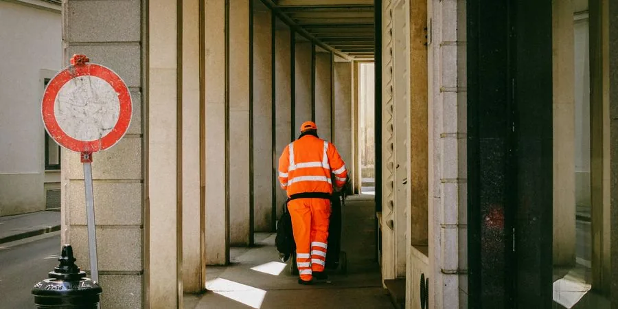 Street worker in orange uniform pushing a cart through a narrow urban alley — the tight-access conditions a self-loading mini dumper replaces at 5–10 times the throughput