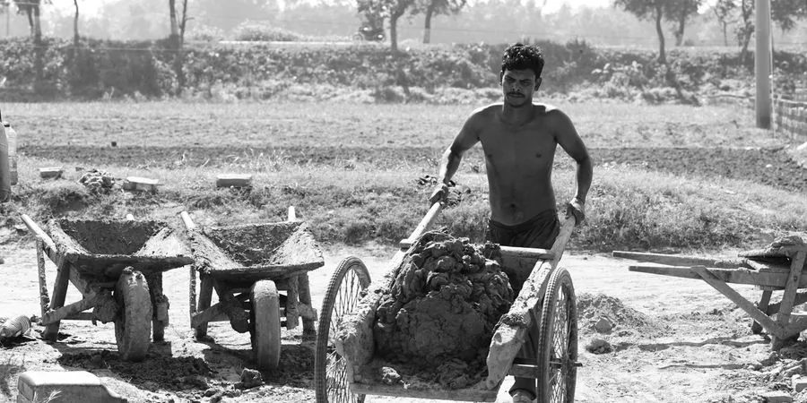 Worker pushing a heavily loaded wheelbarrow full of rocks across a rural site — the manual alternative a motorised power wheelbarrow replaces
