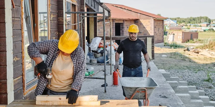 Construction workers moving building materials by hand on a residential site — the manual process a power wheelbarrow replaces